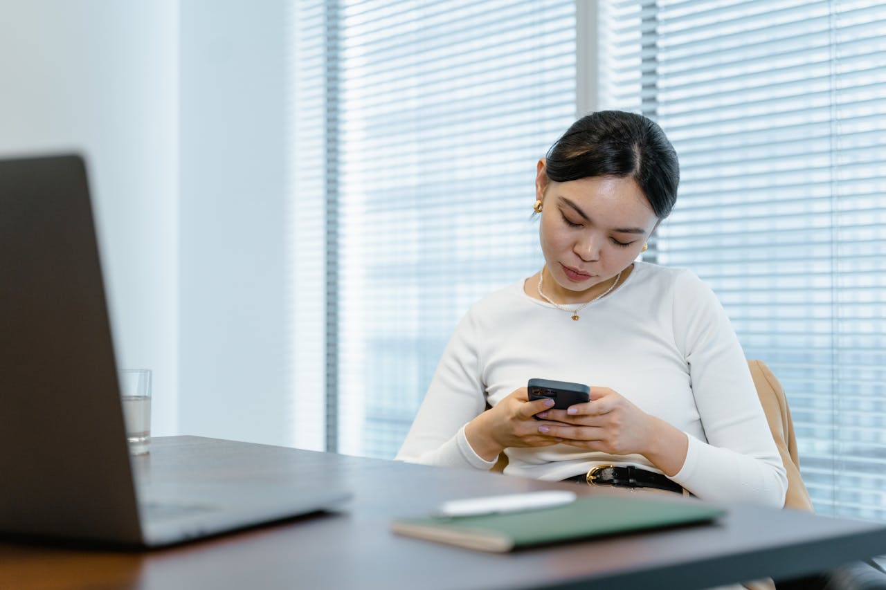 The Art of Drawing Readers In: Your attractive post title goes here Asian woman sitting at her desk, texting on a smartphone in a modern office setting.